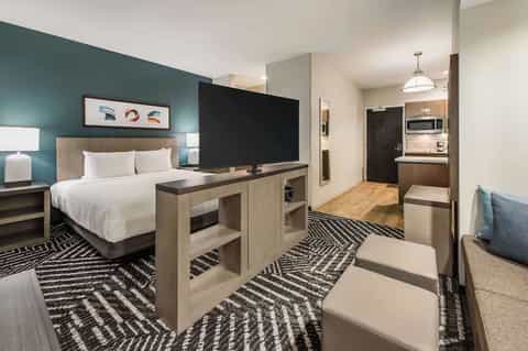 Modern hotel suite with queen bed, teal accent wall, flat-screen TV, kitchenette, and geometric patterned carpet