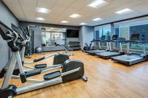 Bright fitness center with exercise bikes, treadmills, weight equipment, and city views through large windows