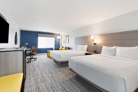 Modern hotel room with two queen beds, blue accent wall, work desk, and bright windows