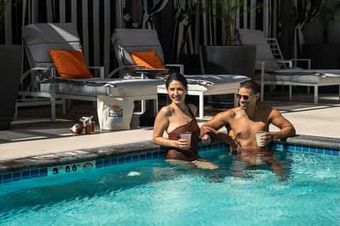 Two guests enjoying drinks in resort pool with loungers and striped umbrellas poolside