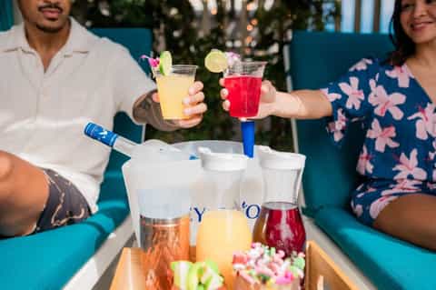 Couple holding colorful cocktails by pool with fruit pitcher and garnishes on blue lounge chairs