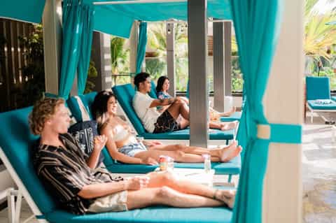 Guests lounging on turquoise poolside chairs under teal pergola with curtains and modern architecture