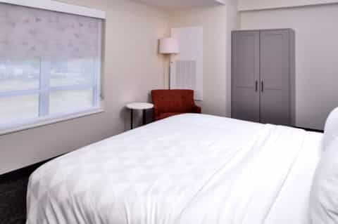 Modern hotel bedroom with queen bed, window, chair, lamp, and closet in neutral tones