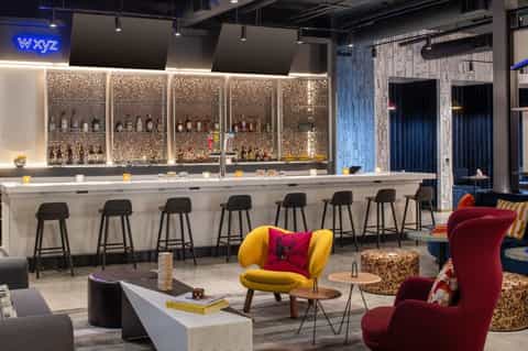 Modern hotel bar with illuminated bottle display, yellow and burgundy accent chairs, and contemporary lounge seating