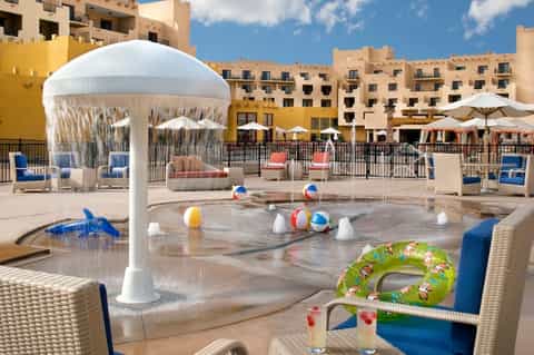 Kids' splash pad with white umbrella structure, colorful beach balls, and resort buildings in background