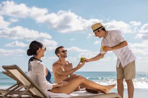 Server in white shirt and hat bringing drinks to couple relaxing on beach loungers