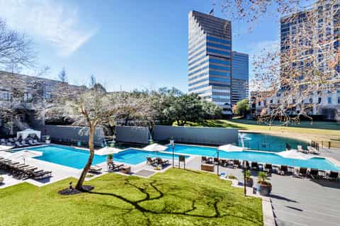 Modern resort pool with white umbrellas, green lawn, and high-rise hotel towers overlooking the deck
