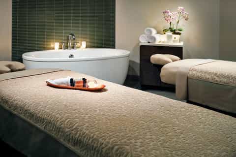 Luxury spa room with soaking tub, massage bed, green tile wall, and fresh flowers