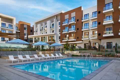 Resort swimming pool with blue umbrellas, loungers, and multi-story brick building complex