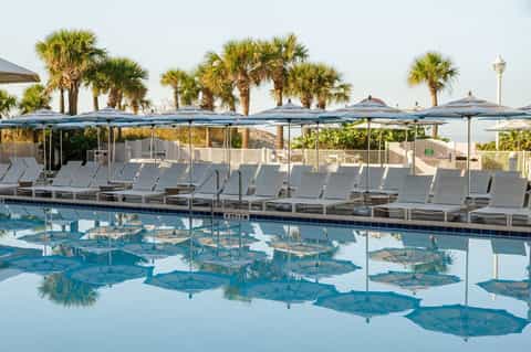 Resort pool with blue umbrellas and lounge chairs, palm trees in background, clear water reflection