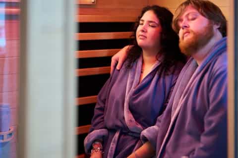 Two guests in robes relaxing together inside infrared sauna with ambient red light