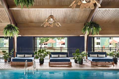 Indoor pool with three wooden lounge beds, hanging plants, and woven light fixtures under wooden ceiling.