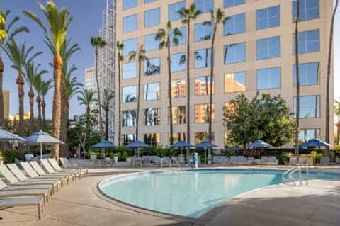 Resort pool with blue umbrellas and lounge chairs, palm trees, and modern high-rise hotel building in background