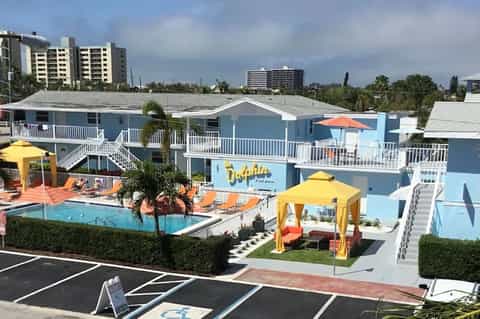 Bright blue motel with pool, yellow umbrellas, white railings, and palm trees from aerial view.