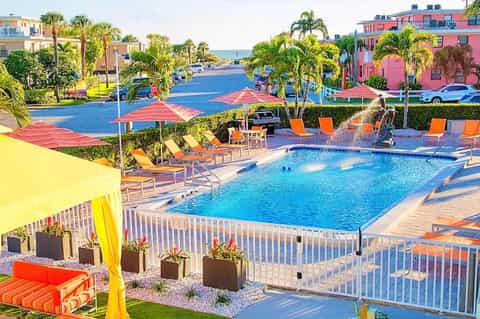 Colorful resort pool area with orange lounge chairs, palm trees, and pink waterfront buildings in background