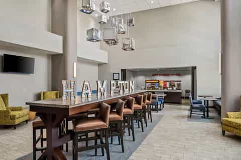 Hotel lobby and breakfast area with high ceilings, HAMPTON sign, bar seating, and modern décor