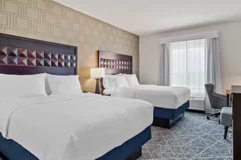 Hotel double queen room with white bedding, blue bed base, patterned wallpaper, and balcony access