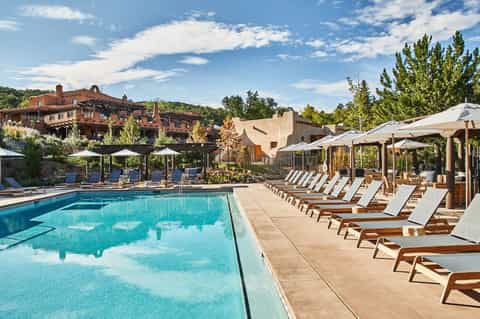 Resort swimming pool with wooden loungers, white umbrellas, and terracotta buildings in background