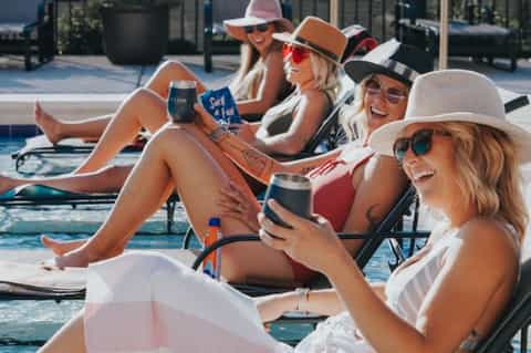 Group of women relaxing by a resort pool wearing cowboy hats and summer attire, holding drinks and enjoying poolside activities