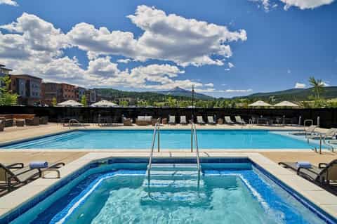 Resort pool area with mountain views, lounge chairs, umbrellas, and spa pool under blue sky