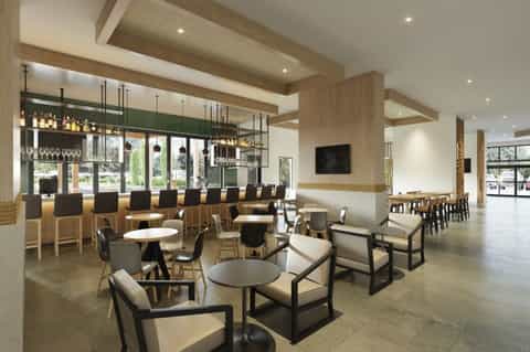 Modern hotel bar and lounge with wooden ceiling beams, green accent wall, and casual seating areas with round tables