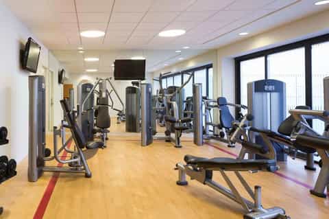 Bright fitness facility with exercise machines, stationary bikes, weights, and television monitors on walls