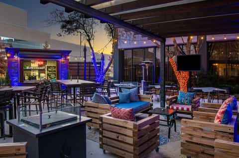 Modern rooftop lounge at dusk with wooden platforms, blue lighting, fire pit, and bar window