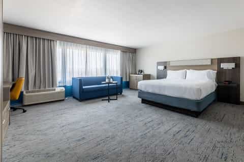 Spacious hotel suite with king bed, blue sofa seating area, floor-to-ceiling windows, and modern wood accents
