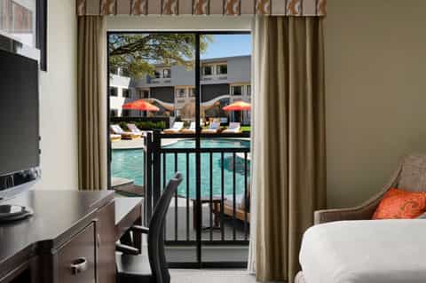 Hotel room with desk, bed, and balcony overlooking pool area with umbrellas and lounge chairs