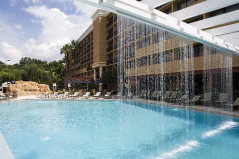 Resort pool with cascading water wall feature, lounge chairs, and multi-story hotel building under blue sky