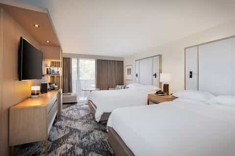 Contemporary hotel room with two queen beds, wood accents, flat-screen TV, and floor-to-ceiling windows with sheer curtains