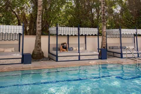 Luxury cabana beds with white striped canopies along a resort pool surrounded by mature trees