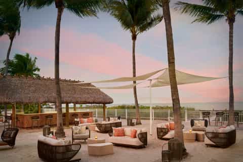 Beachfront lounge area with woven seating, fire pit, and sail shade at sunset with palms