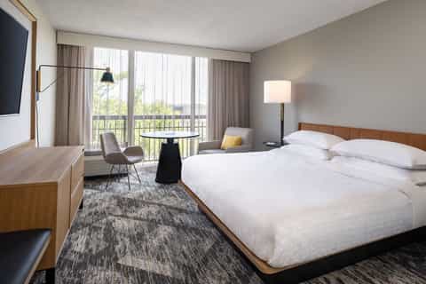 Hotel bedroom with king bed, wooden headboard, balcony access, work desk, and contemporary neutral décor