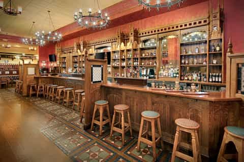 Upscale bar interior with ornate wood cabinetry, red walls, decorative chandeliers, and extensive liquor bottle display