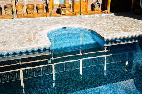 Kidney-shaped pool with blue mosaic tiles, stone decking, and wooden chairs beside building