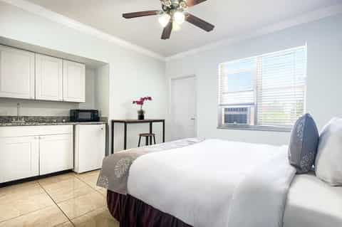Modern hotel room with queen bed, kitchenette, ceiling fan, and large window with shutters