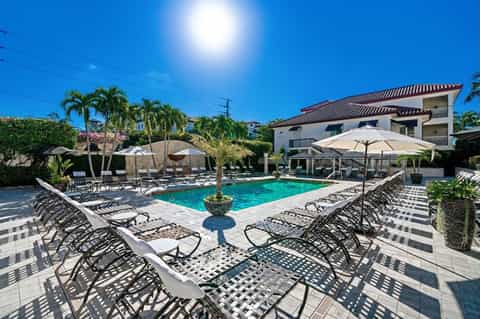 Resort pool surrounded by loungers with umbrellas, palm trees, and multi-story white buildings under blue sky