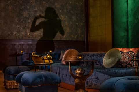 Upscale lounge with jewel-toned velvet seating, brass accents, patterned pillows, and moody atmospheric lighting