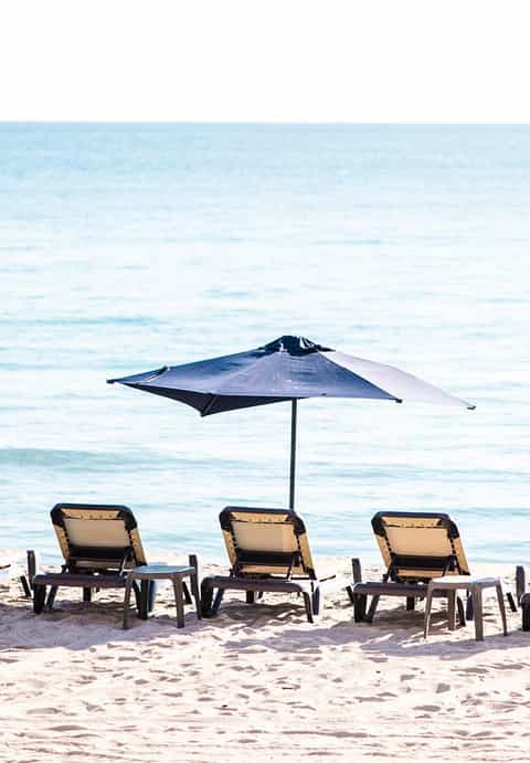 Three lounge chairs with single blue umbrella on sandy beach facing calm turquoise water