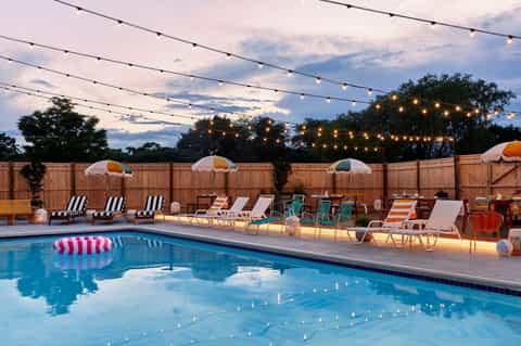 Resort pool at dusk with string lights, umbrellas, lounge chairs, and wooden fence backdrop