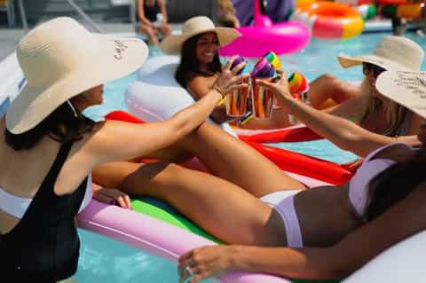 Group of women in sun hats on colorful pool floats clinking rainbow-colored drinks together