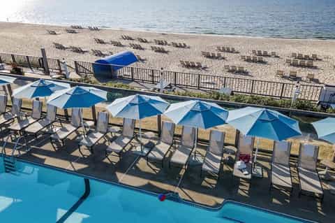 Beachfront pool area with blue umbrellas, lounge chairs, and sandy beach with sunbathers