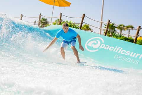 Surfer riding artificial wave on shallow water ride with palm trees and yellow umbrella