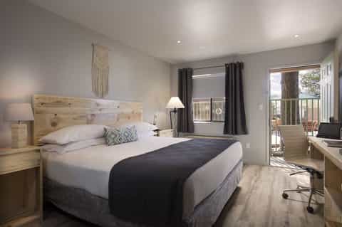 Modern hotel bedroom with queen bed, wooden headboard, desk, and glass door opening to balcony
