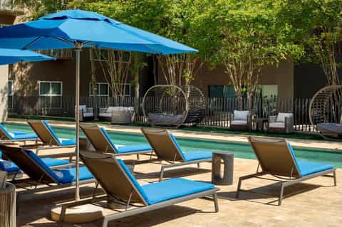 Pool deck with blue loungers, blue umbrellas, hanging egg chairs, and multi-story tan building