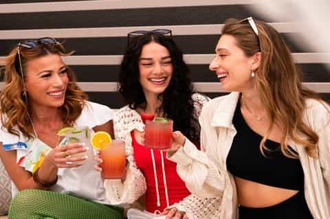 Three women enjoying colorful cocktails by the pool with striped background
