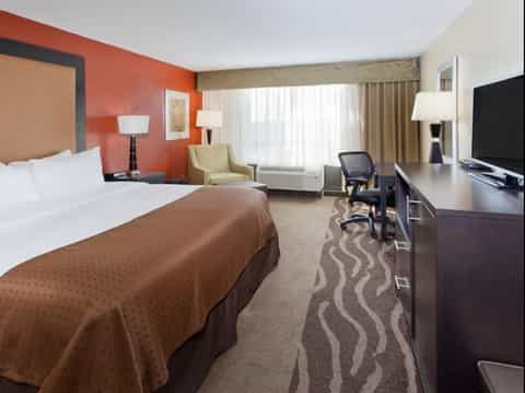 Spacious hotel room with queen bed, orange accent wall, seating area, and flat-screen TV