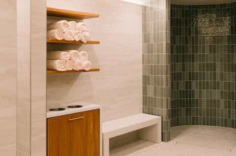 Spa bathroom with rolled white towels on wooden shelves, green tile shower area, and wooden vanity with white countertop