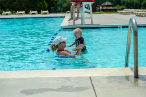 Swim instructor teaching young child in resort pool with diving board and slide visible